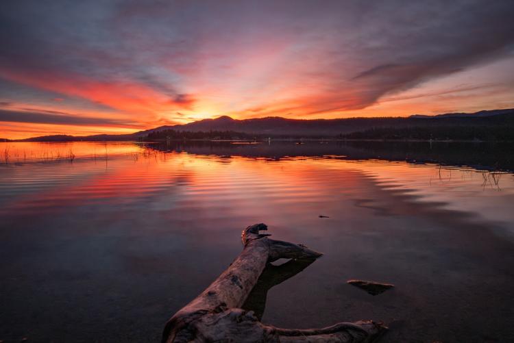 A photo of a bright red-ish pink and orange sunset peering from over the tall mountains, the lake reflecting it like a mirror