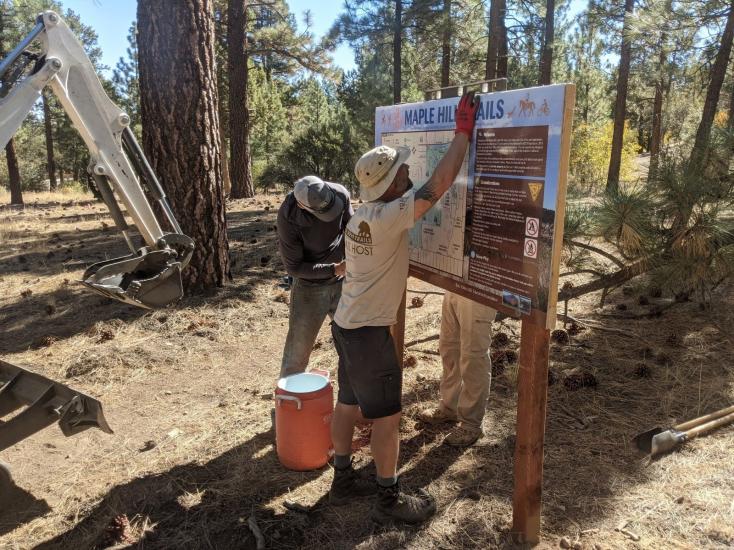 A photo of three people maintaining the Maple Hills Trail sign, there is a excavator scoop next to the tree behind them.